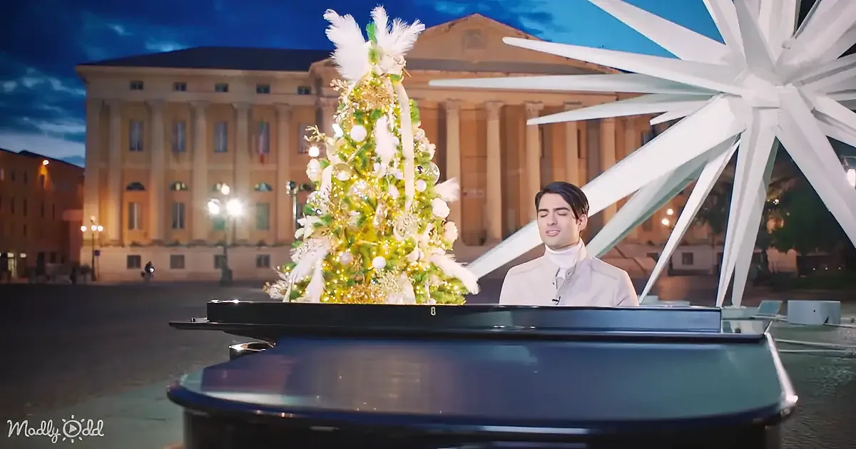 A man in a white coat plays a grand piano in front of a decorated Christmas tree and a large white star, with a grand...