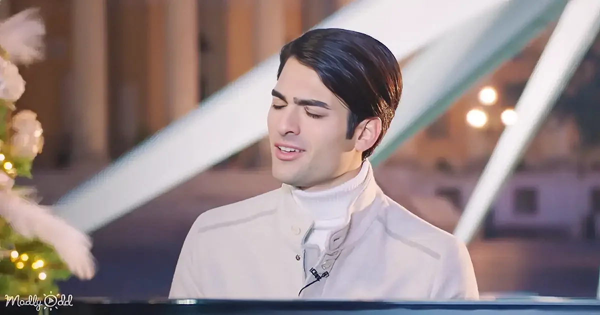 A man in a white jacket and turtleneck sings at a piano, with a Christmas tree and lights in the background.