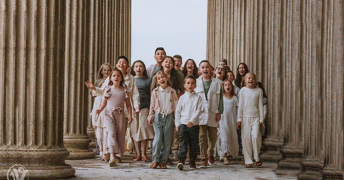 A group of children in white outfits stands between large stone columns, smiling and posing together.