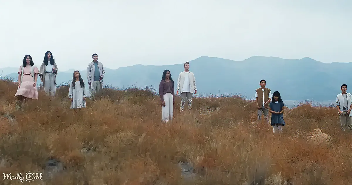 A group of people stands in a field of dry grass, with mountains in the background under a cloudy sky.