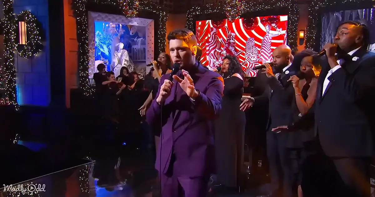 A man in a purple suit sings into a microphone on stage with a group of musicians.