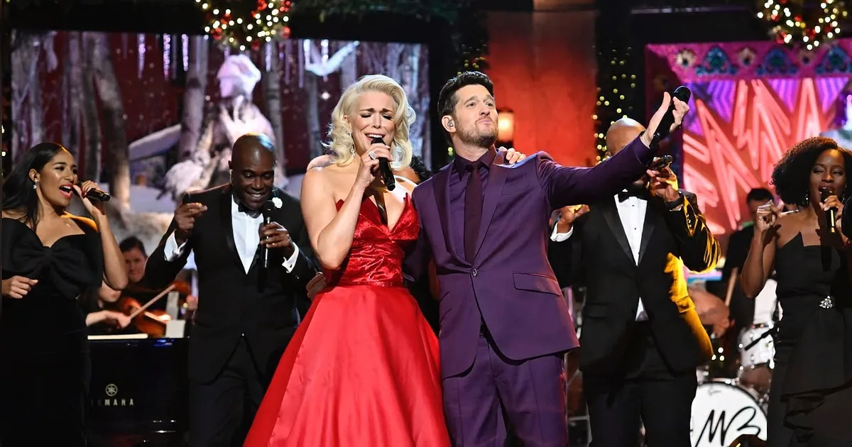 A man in a purple suit and a woman in a red dress sing on stage with a band and other performers.