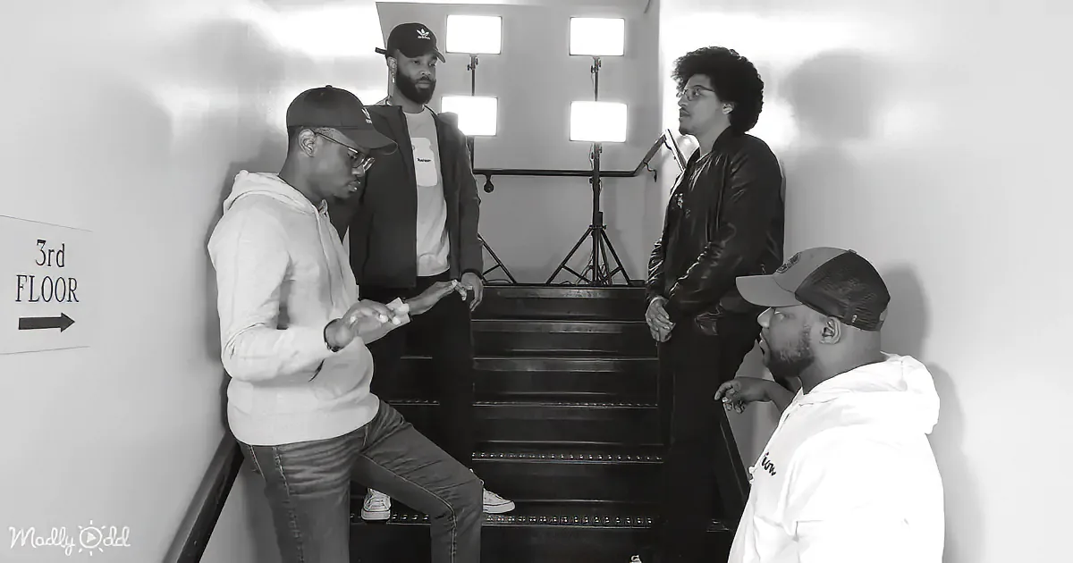 A man in a white hoodie and jeans stands on stairs, holding something, with three others around him.
