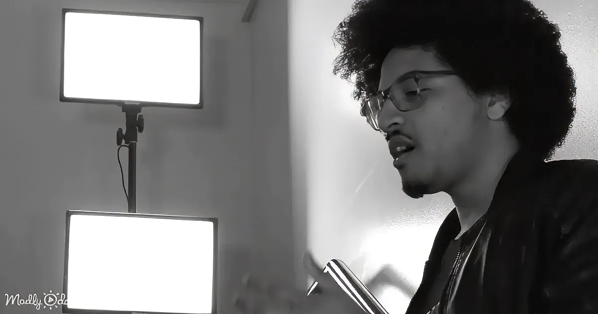 A person with curly hair and glasses is holding a microphone, possibly singing or speaking, in a room with two bright lights.