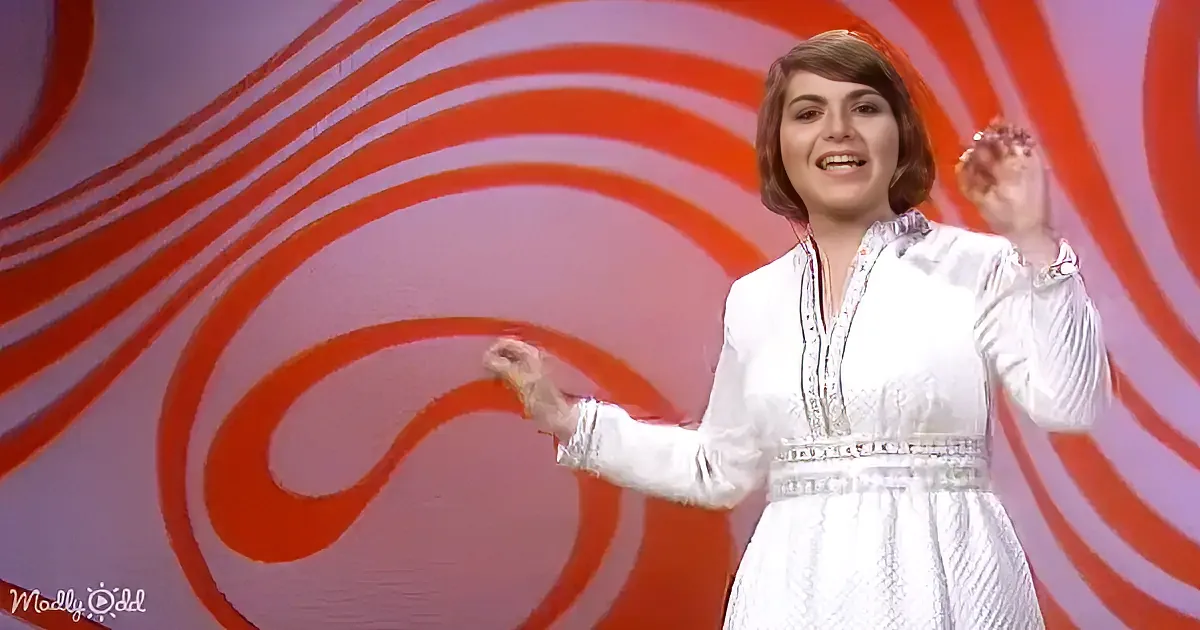 A woman in a white dress with a red and white swirl background smiles and gestures with her hands.