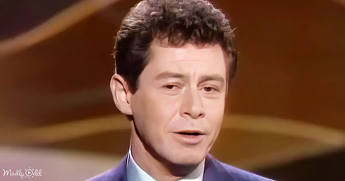 A man with curly hair, wearing a blue suit, is speaking with a serious expression.