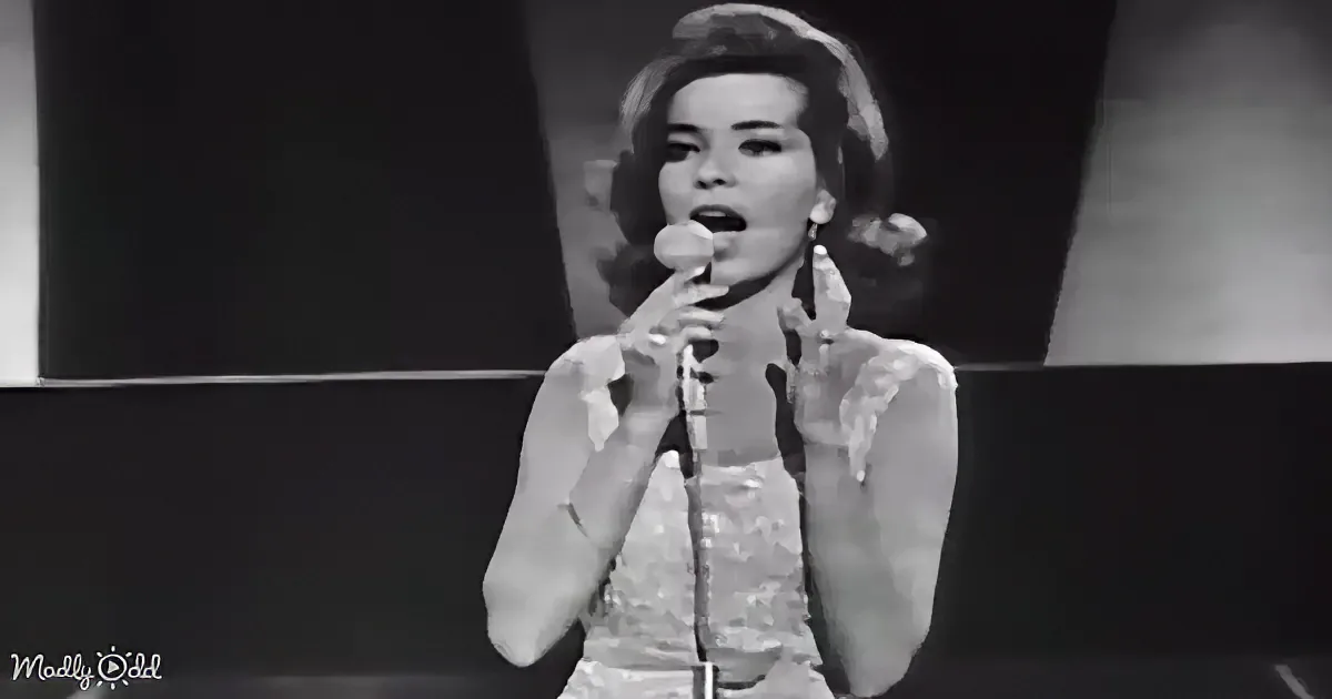 A woman in a white dress sings into a microphone, her hands raised, in a black-and-white image.