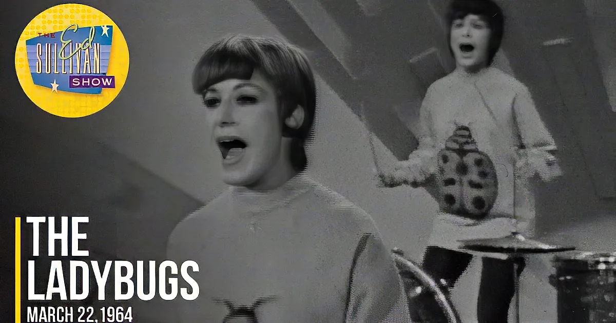 A woman in a beige turtleneck sweater with a ladybug design smiles in front of a drum set.