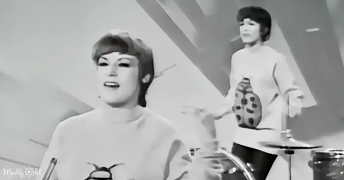 A woman in a white top with a ladybug design sings in the foreground, another woman plays drums in the background.