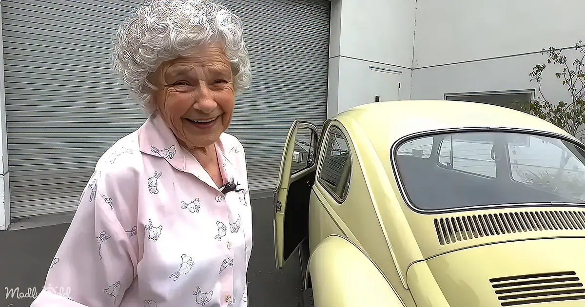 An elderly woman in a pink pajama top smiles near a vintage yellow Volkswagen Beetle with its door open.