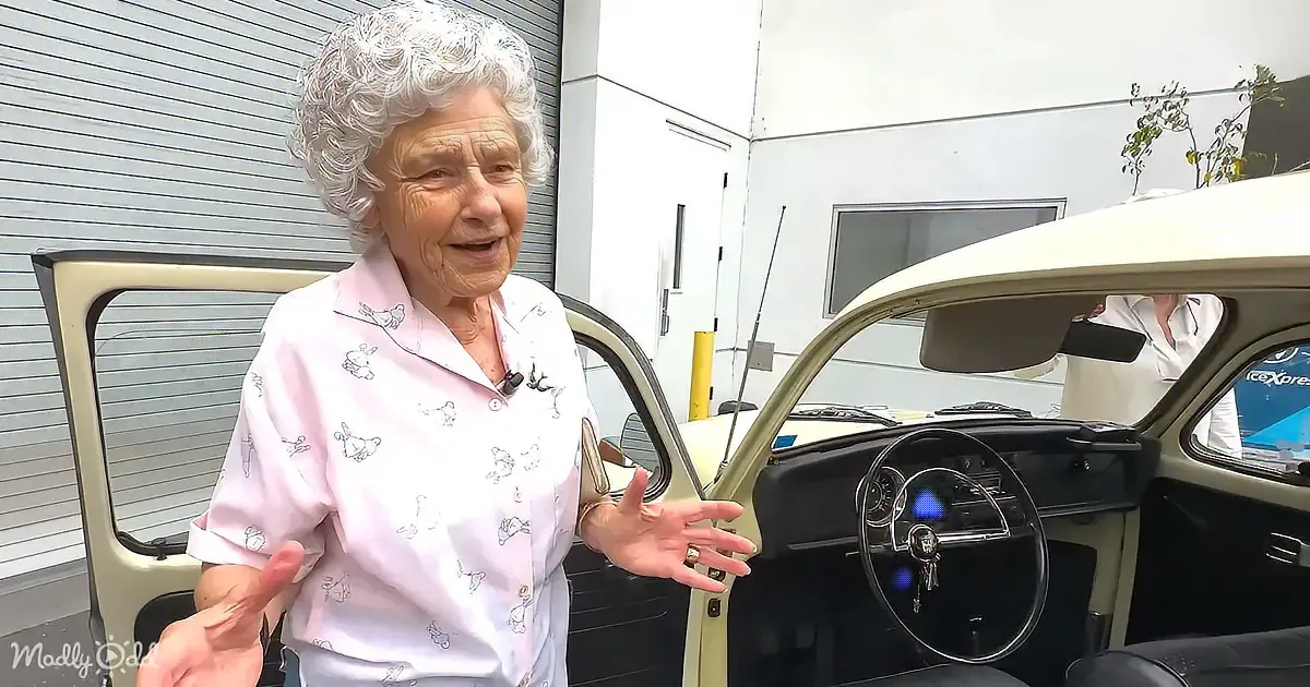 An elderly woman in a pink shirt stands next to a vintage car, gesturing with her hands.