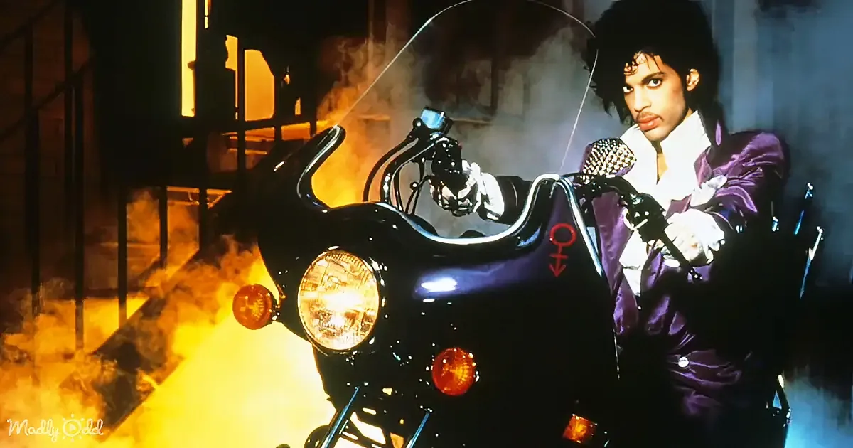 Prince in a purple suit rides a black motorcycle with a glowing headlight amidst smoke and fire.