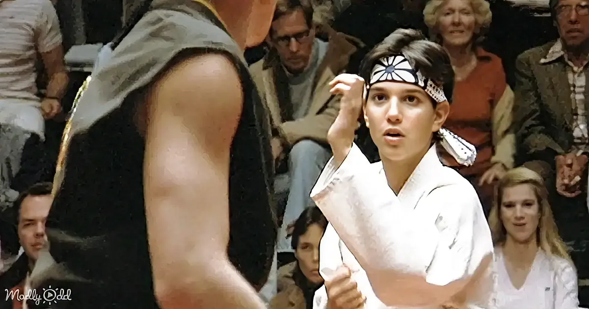 A woman in a white karate uniform with a headband stands in front of a man in a black vest, surrounded by spectators.