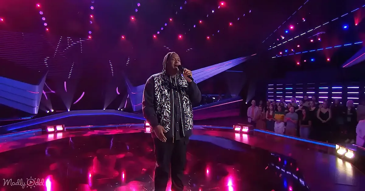 A man in a hoodie sings into a microphone on a stage with colorful lights and an audience.