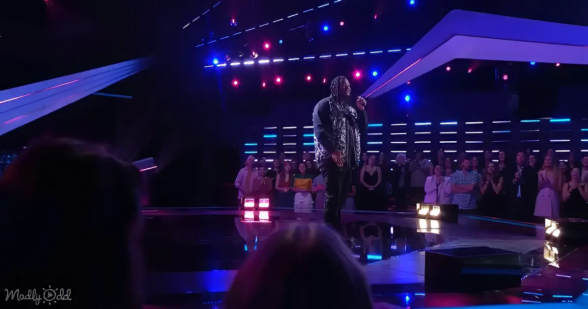 A person in a patterned outfit sings on stage with bright lights and an audience clapping.