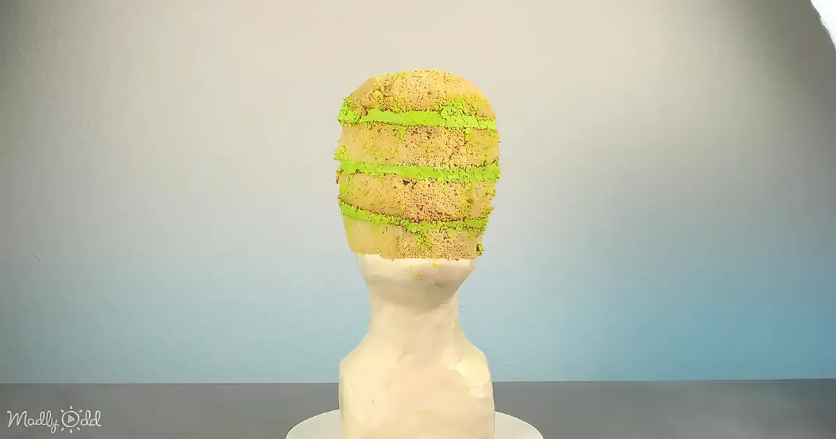 A cake resembling a head with green frosting lines sits on a mannequin bust against a plain background.