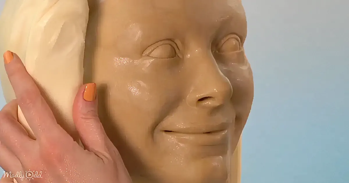 A hand with orange nail polish touches a smooth, beige-colored face sculpture against a light blue background.