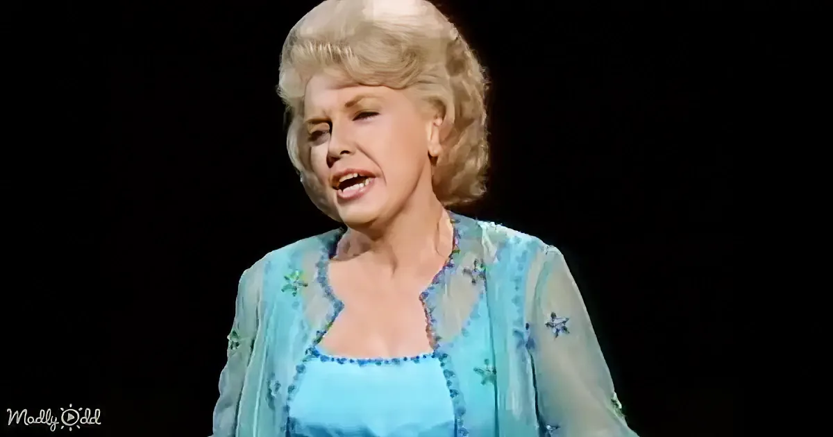 A woman with blonde hair styled in a 1950s fashion, wearing a blue dress with floral patterns, is shown against a black...