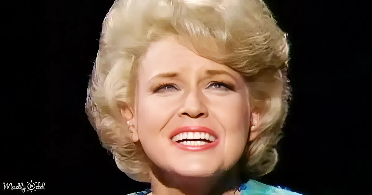 A woman with blonde, voluminous hair and red lipstick is smiling, wearing a blue patterned top.