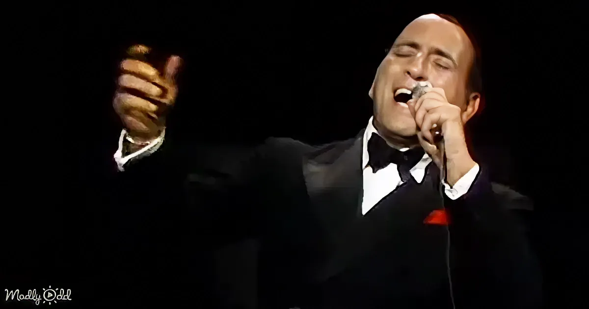 A man in a tuxedo sings passionately into a microphone, raising his hand in a gesture of enthusiasm.