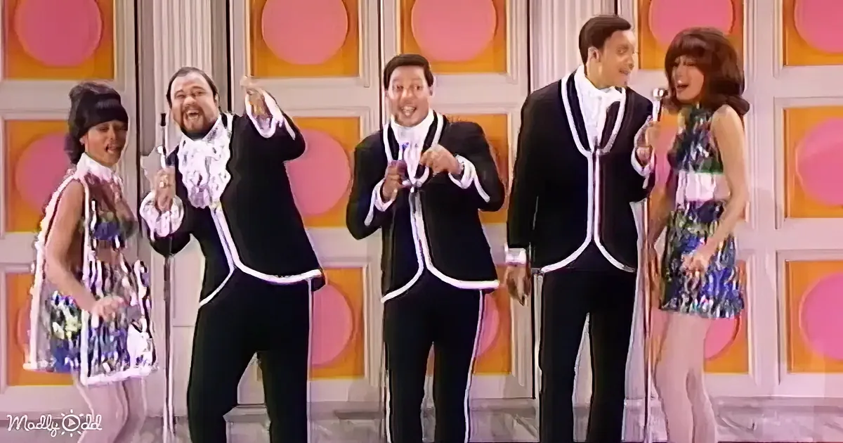 A group of performers in black suits with white trim sing on stage, one holding a microphone.