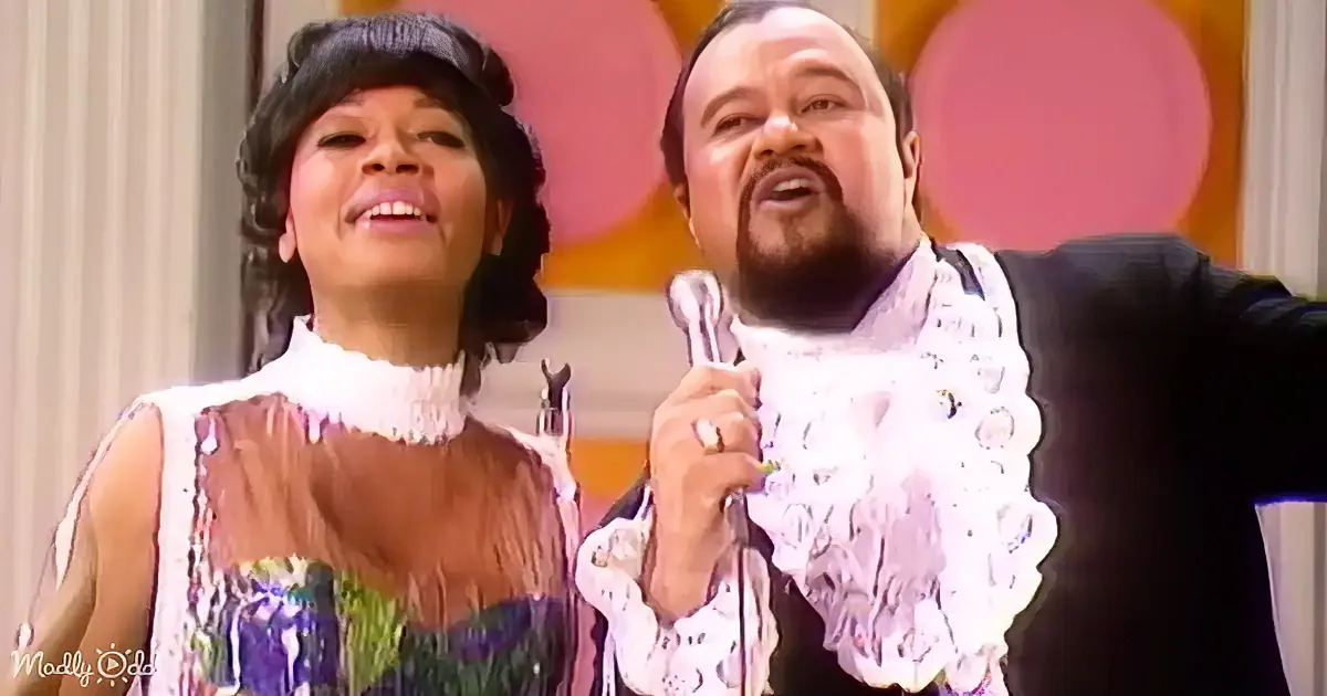 A man in a white ruffled shirt holds a microphone, standing next to a woman in a floral dress, both smiling.