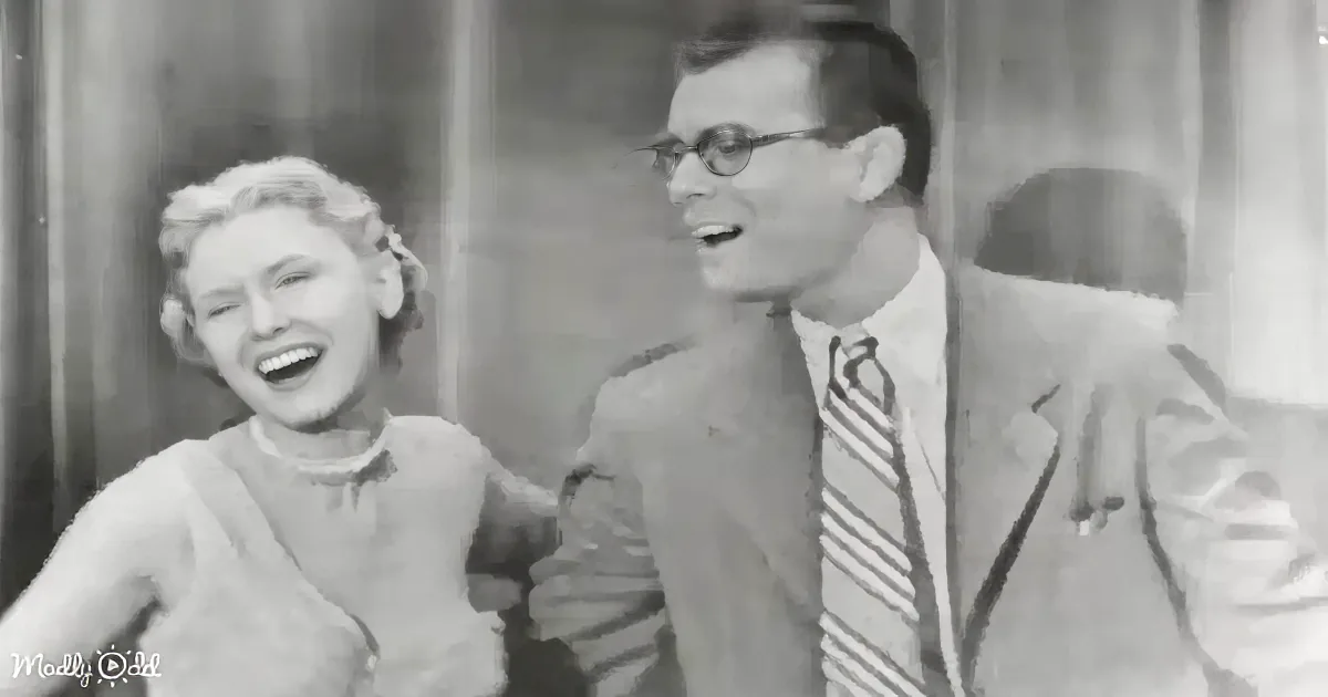 A man in a suit and striped tie sits beside a woman laughing, both appear to be in a studio setting.