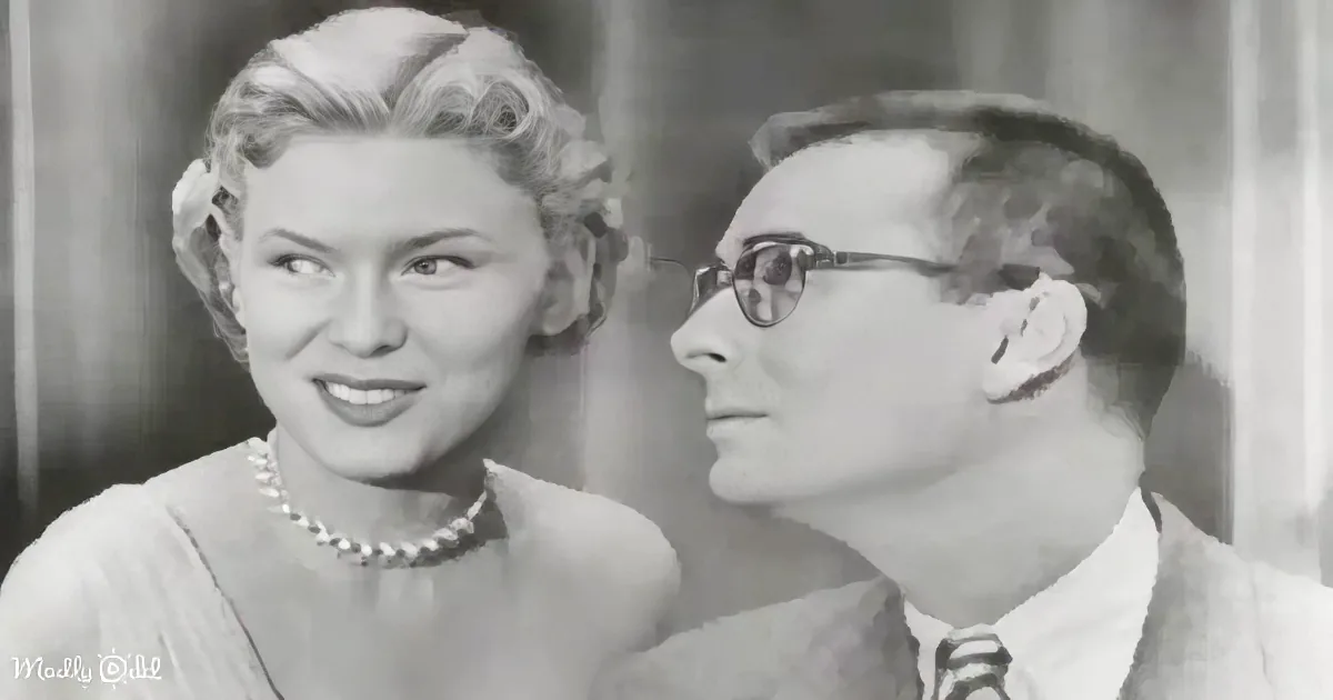 A woman in a pearl necklace smiles while a man in glasses looks on, both in formal attire.