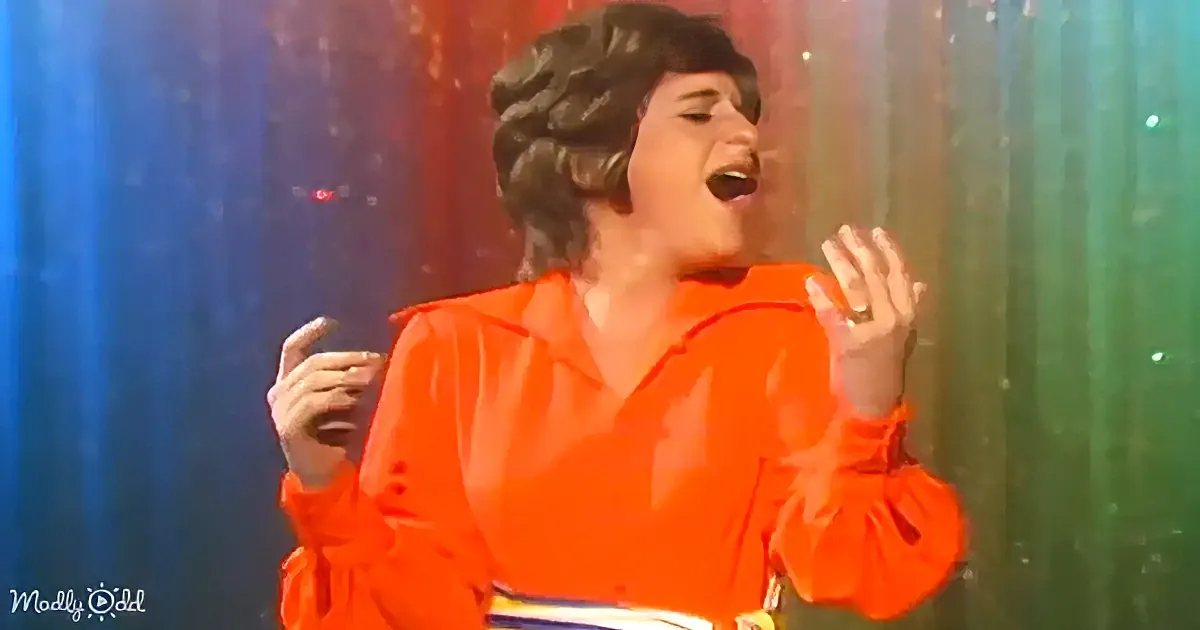 A person in a bright orange outfit sings passionately with hands raised, against a colorful, glittery background.