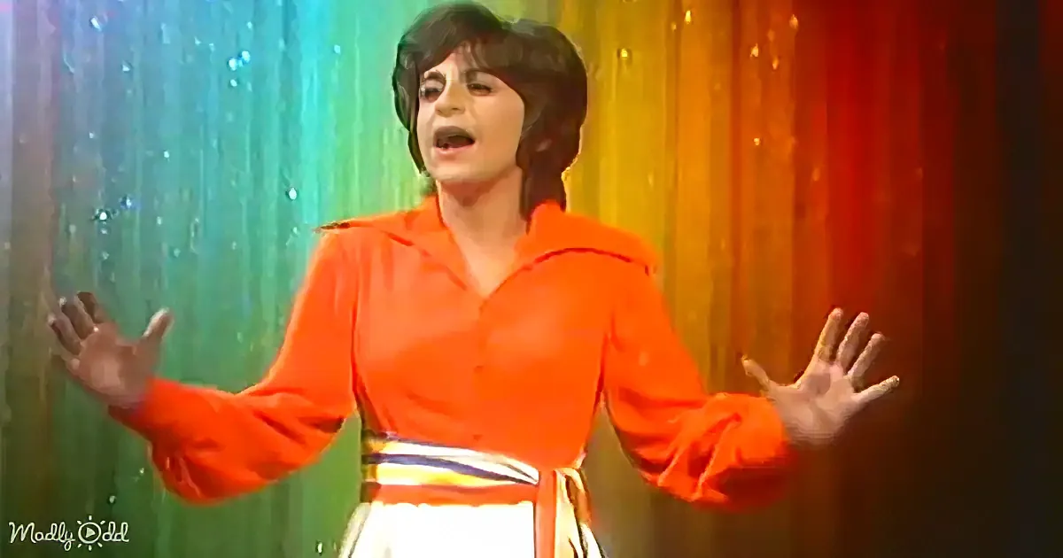 A person in a vibrant orange shirt and white skirt stands with arms outstretched against a colorful, glittery backdrop.