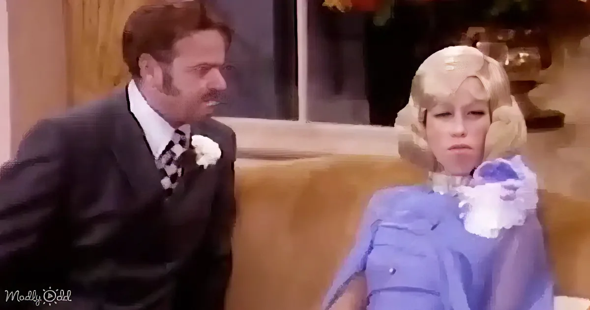 A man in a suit and a woman in a blue dress with a white flower on her lapel are seated on a couch.