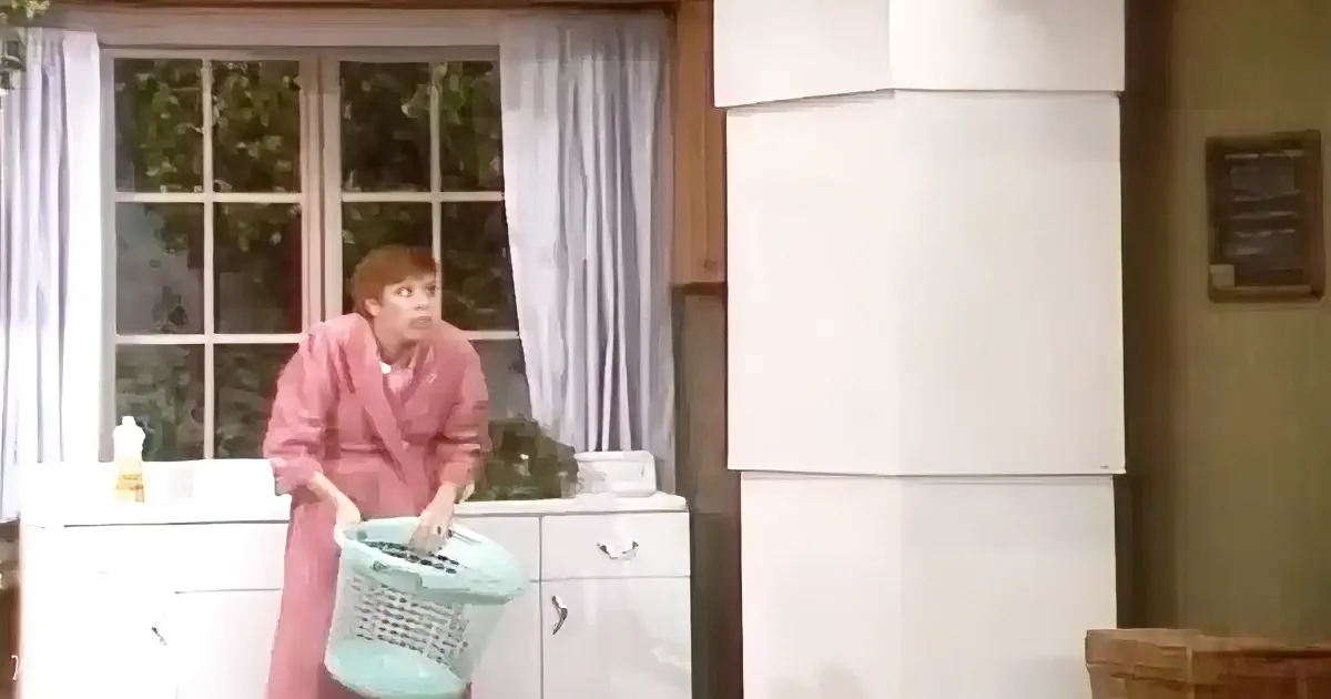Woman in pink robe holds laundry basket, stands in kitchen near white cabinets and window.