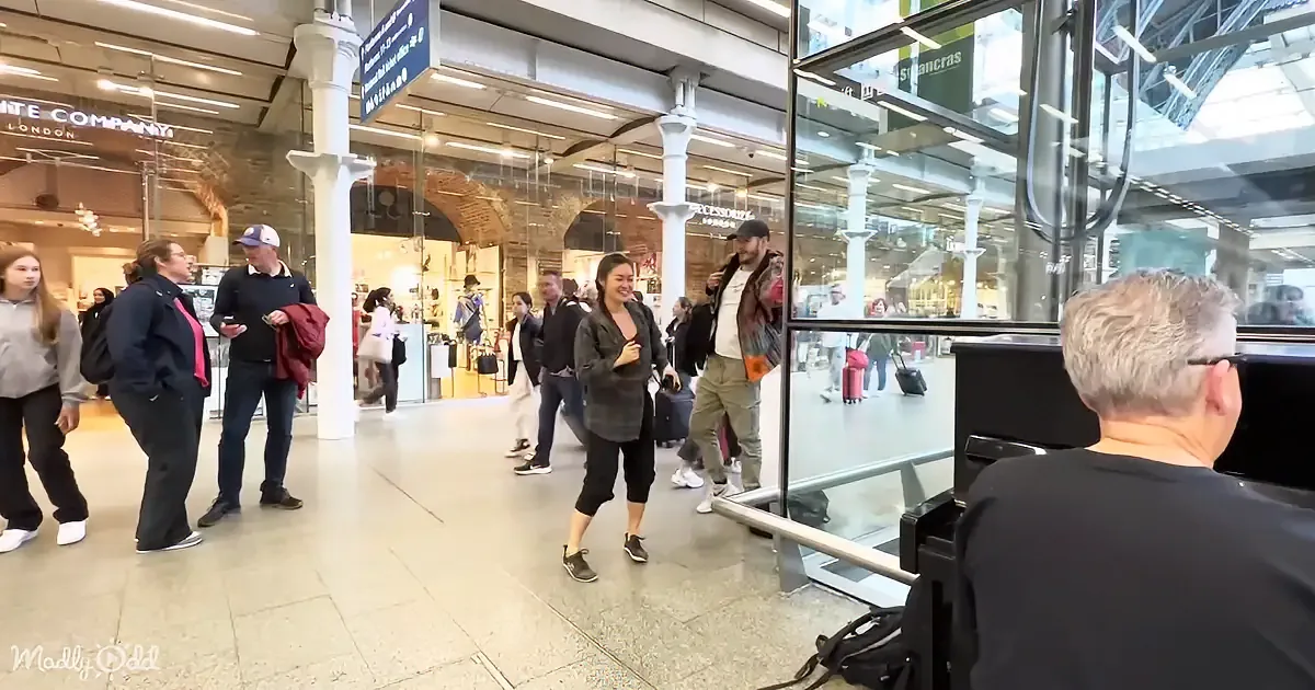A man with a camera films a group of people walking in a busy shopping area with stores and glass windows.