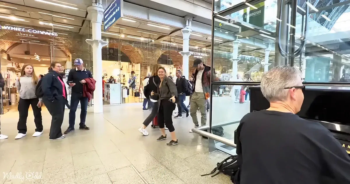 A woman in a black jacket and jeans walks through a busy train station with luggage.