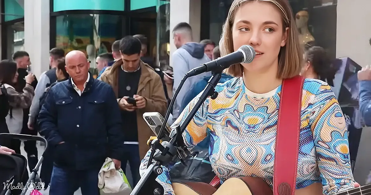A woman in a colorful patterned shirt sings into a microphone while playing guitar, surrounded by a busy street scene.