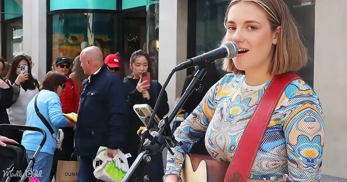 A woman in a colorful patterned shirt sings into a microphone while holding a guitar, with a crowd of people in the...