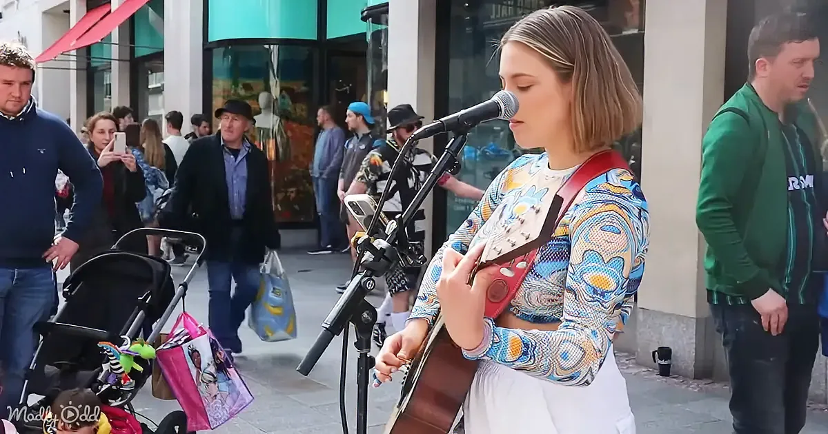 A woman plays guitar and sings into a microphone on a busy street, surrounded by pedestrians.