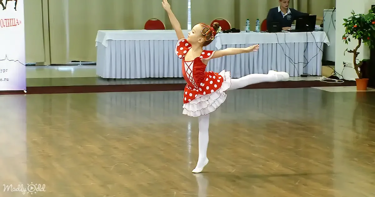 A young girl in a red polka-dot ballet costume performs a dance move on a wooden floor.