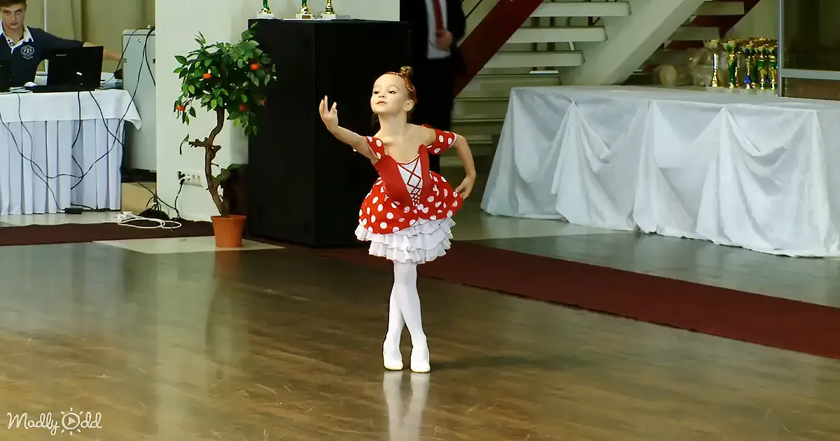 A young girl in a red polka-dot ballet costume dances gracefully on a wooden floor.