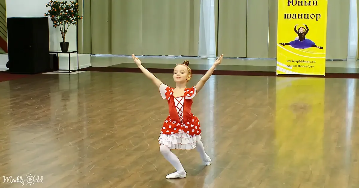 A young girl in a red polka-dot ballet outfit dances gracefully on a wooden floor.