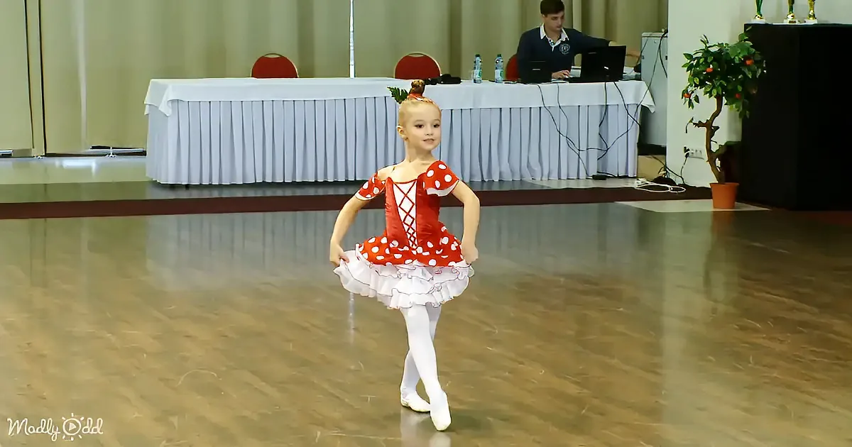A young girl in a red polka-dot ballet outfit stands on a wooden floor, holding her skirt.