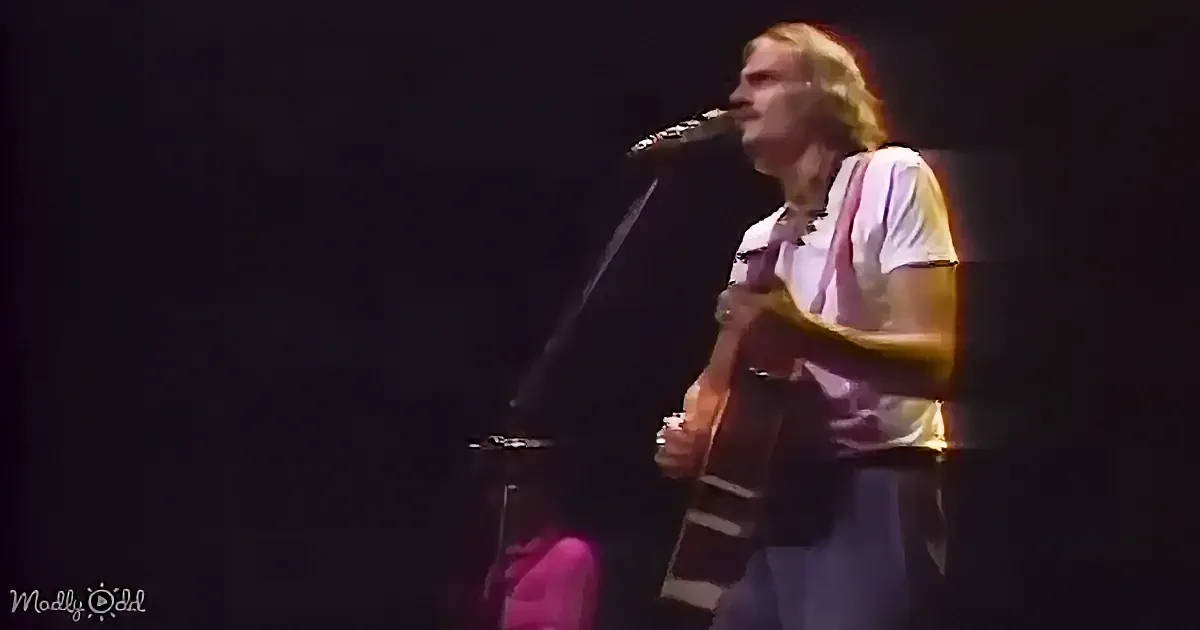 A man with long hair sings into a microphone while playing an acoustic guitar on stage.