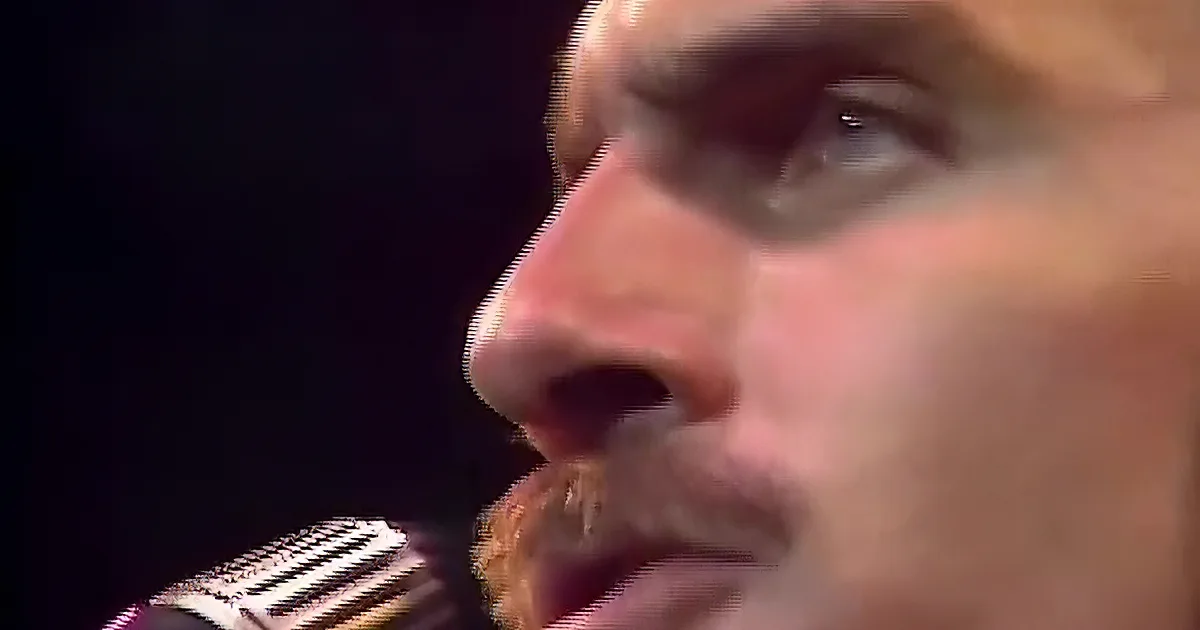 A man with long, wavy hair sings into a microphone, wearing a white shirt with red stripes.