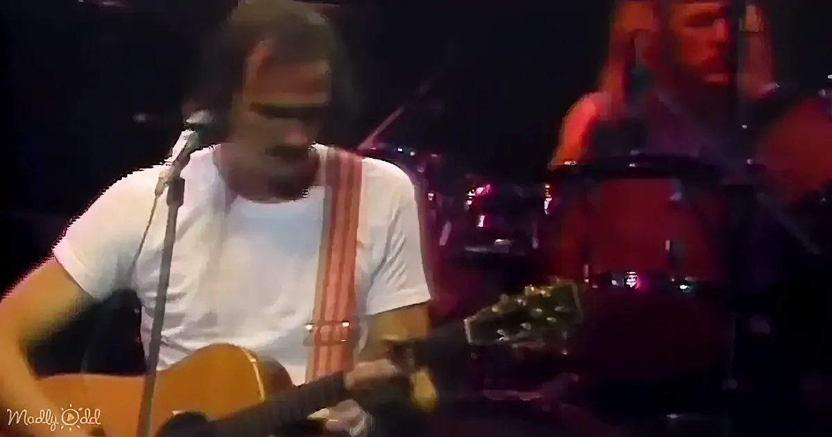 A man in a white shirt plays an acoustic guitar on stage, with a microphone and drum set in the background.