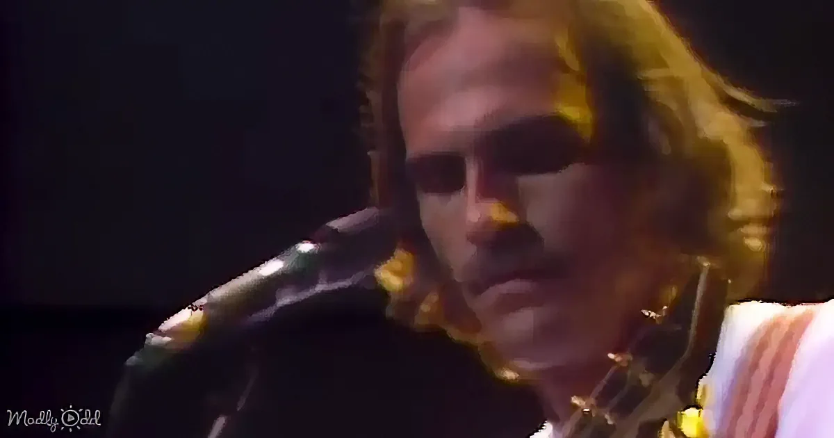 A person with long hair and sunglasses is singing into a microphone, wearing a white shirt with red stripes.