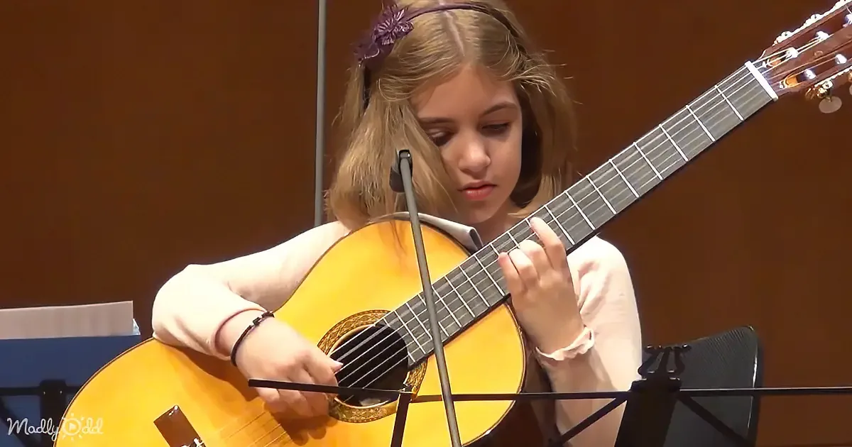 A young girl with a headband plays a classical guitar in front of a microphone.