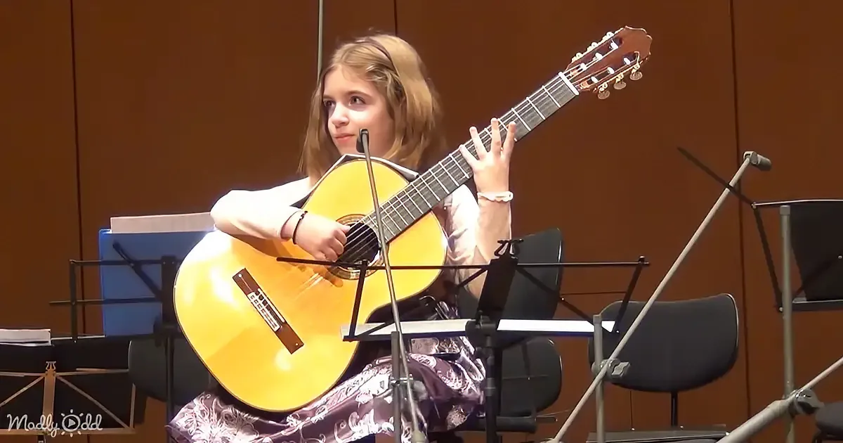 A young girl plays a classical guitar in a music setting with sheet music stands and chairs.