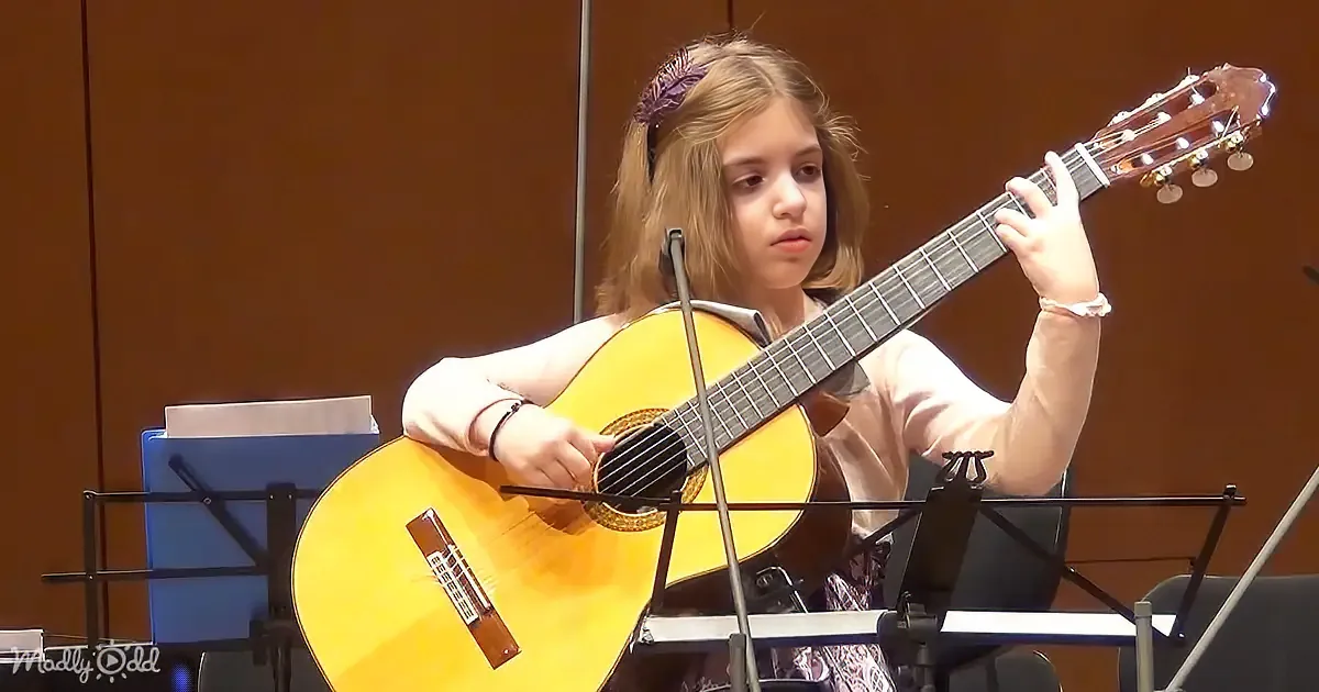 A young girl plays a classical guitar in front of a music stand.
