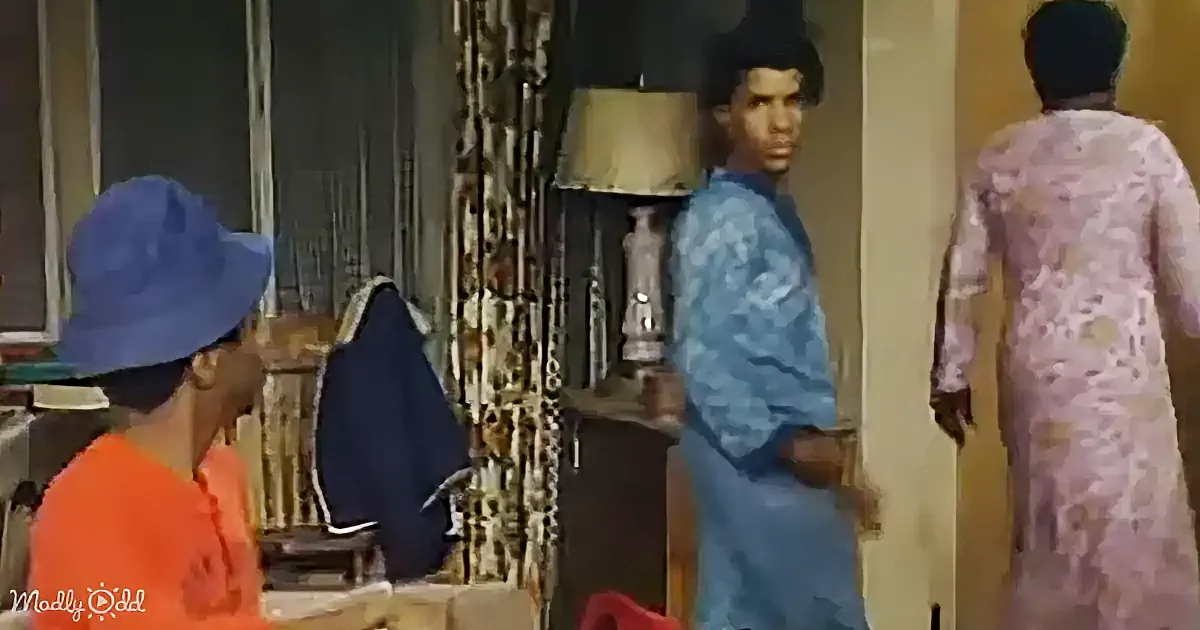A man in a blue outfit stands in a room with a lamp and patterned curtains, while another person in an orange shirt and...