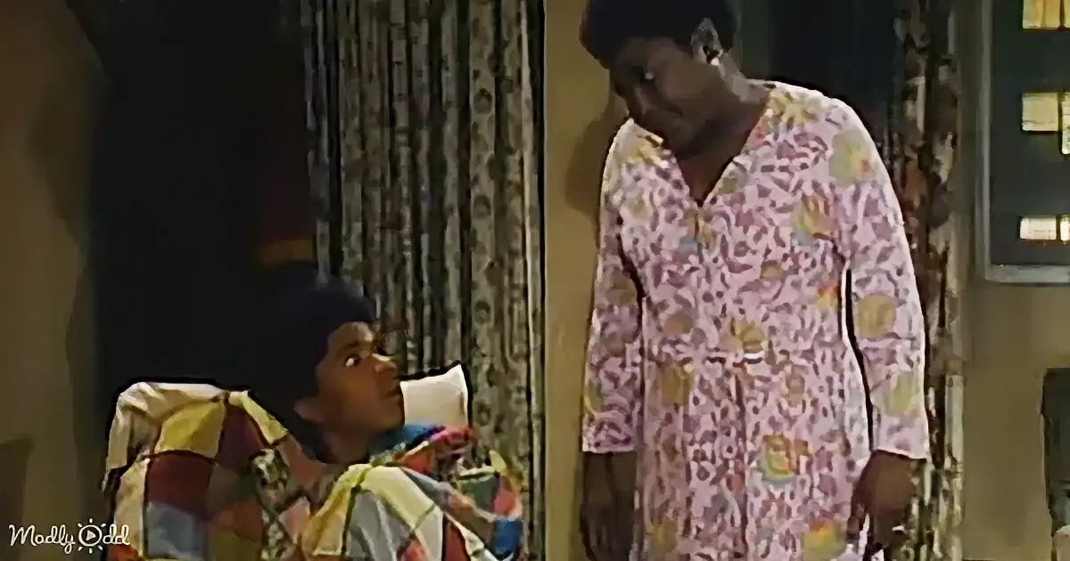 A person in pajamas stands near another person in a colorful bed, both in a room with curtains and a window.
